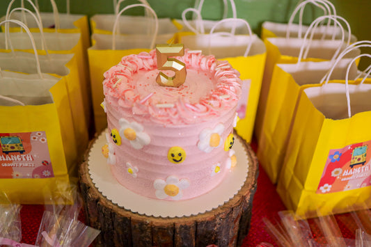 Pink birthday cake surrounded by colorful party favor goodie bags