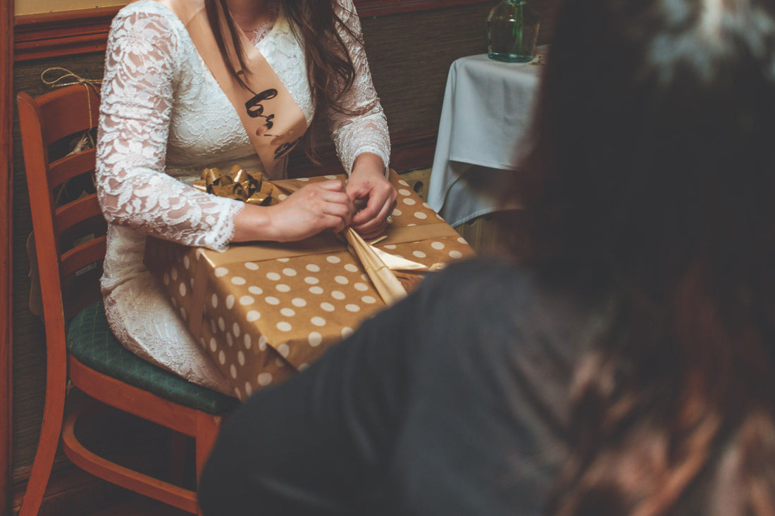 Bridal shower guest of honor in a lace dress and sash unwrapping a wrapped gift
