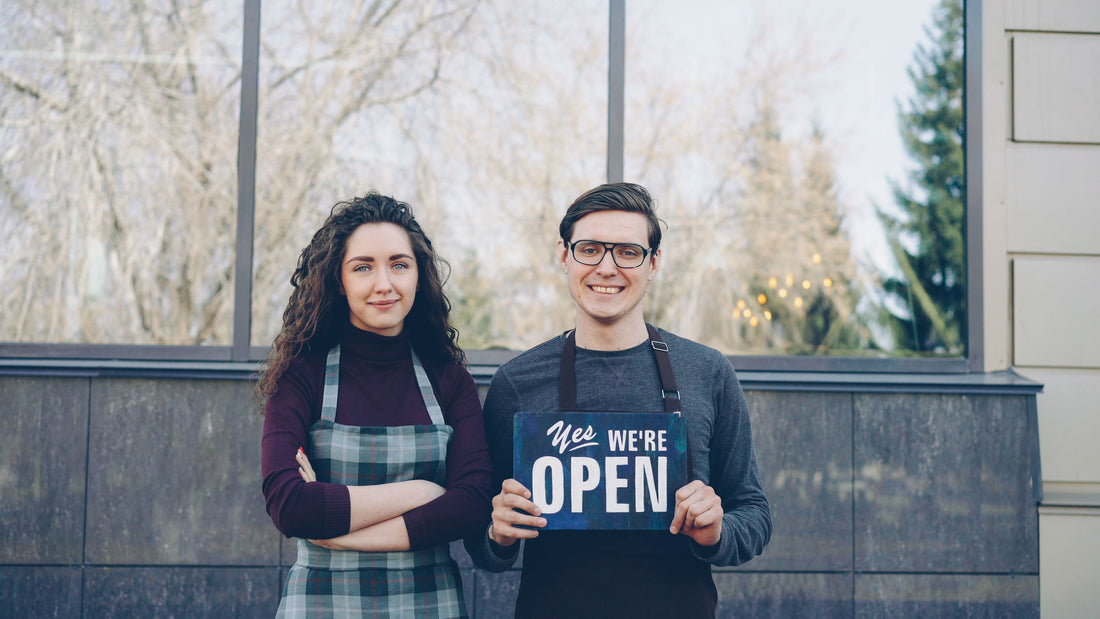 Two welcoming small business owners standing outside their storefront holding an open sign