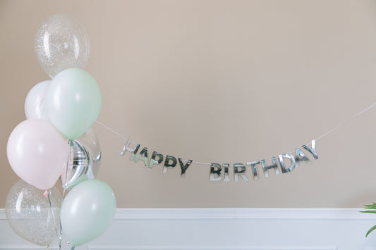 Minimalist pastel balloon bouquet with a silver Happy Birthday banner against a plain wall