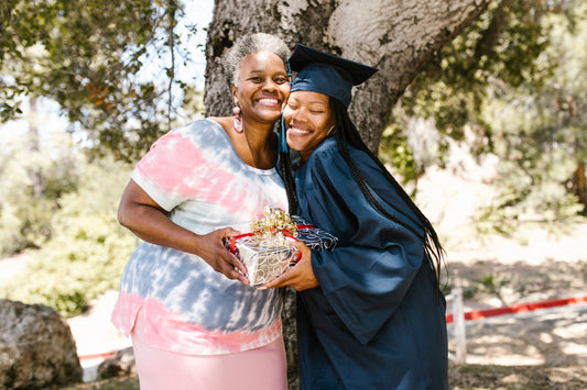 Graduate in cap and gown hugging family member outdoors while celebrating with a gift
